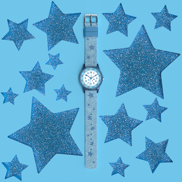 Eine Kinder-Armbanduhr mit einem hellblauen Glitzerarmband, auf dem dunkelblaue Sternchen abgebildet sind, blau-transparentem Gehäuse mit eingearbeitetem Glitzer und kleinen Schmucksteinen auf dem Ziffernblatt. Die Zahlen auf dem blau- weißem Ziffernblatt sind sehr gut ablesbar.