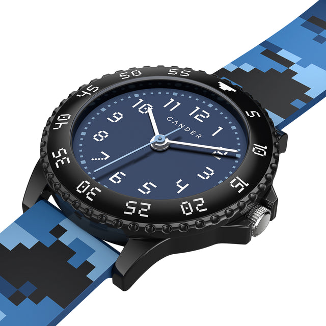 Eine Kinder-Armbanduhr in Pixel-Look. Auf dem Armband sind blaue und schwarze Pixel zufällig angeordnet. Die weißen Ziffern auf dem schwarzem Gehäuse und dem blauen Ziffernblatt sind ebenfalls im Pixel-Look. Die Uhr verfügt über eine verstellbare Lynette in schwarz. 