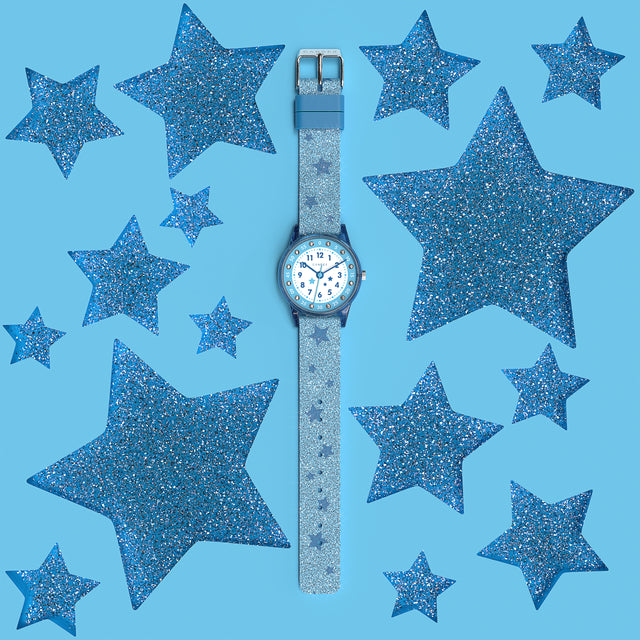 Eine Kinder-Armbanduhr mit einem hellblauen Glitzerarmband, auf dem dunkelblaue Sternchen abgebildet sind, blau-transparentem Gehäuse mit eingearbeitetem Glitzer und kleinen Schmucksteinen auf dem Ziffernblatt. Die Zahlen auf dem blau- weißem Ziffernblatt sind sehr gut ablesbar.