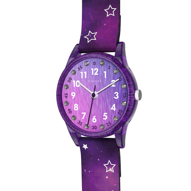 Eine Kinder-Armbanduhr mit einem lila Stoffarmband, auf dem kleine Sterne abgebildet sind. Das lila-transparente Gehäuse hat ein Glitzereffekt. Auf dem Ziffernblatt befinden sich kleine Schmucksteine. Die weißen Zahlen auf dem lila Ziffernblatt sind sehr gut ablesbar.